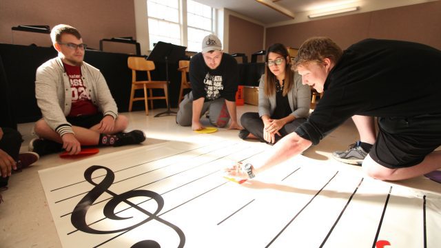 students in a music ed classroom make use of a music staff imprinted rug on the floor