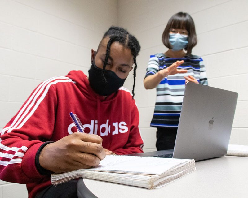 Student writing on a notebook in the background who are both wearing masks