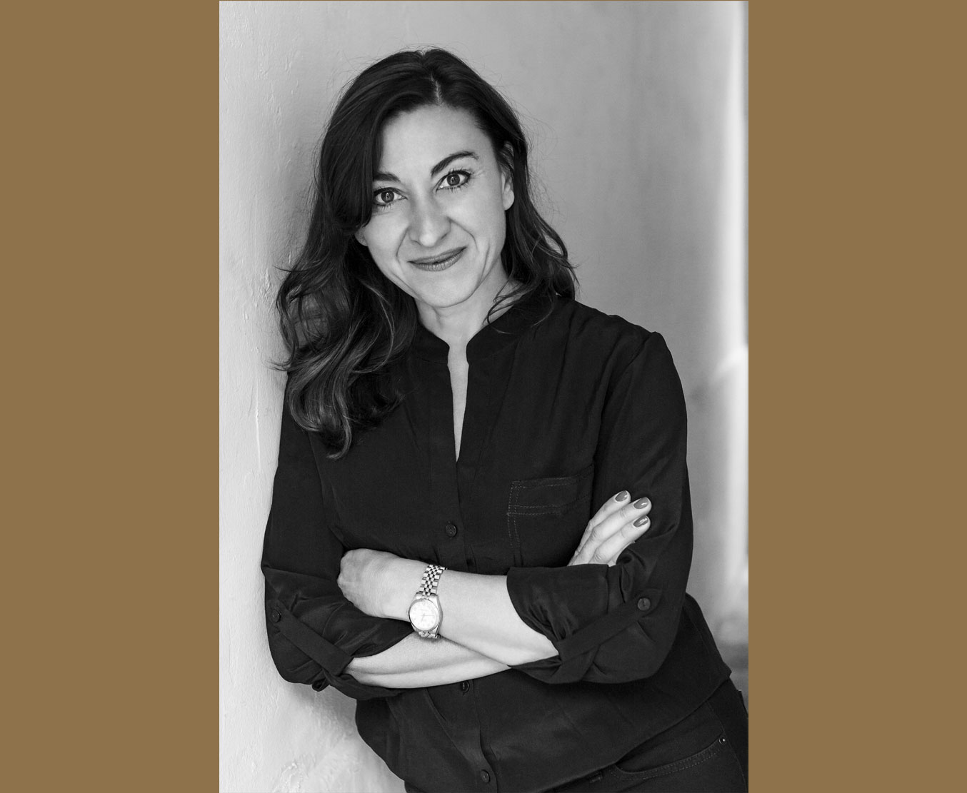 A black and white portrait of Lynsey Addario, who stands with her arms crossed while leaning against a wall and smiling