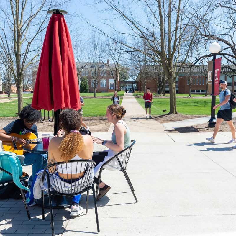 Three students sitting around a umbrella table with more students walking in the background