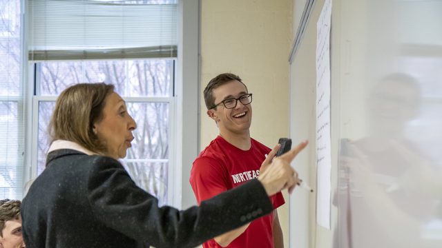 Professor in the foreground pointing to a white board with student smiling in the background