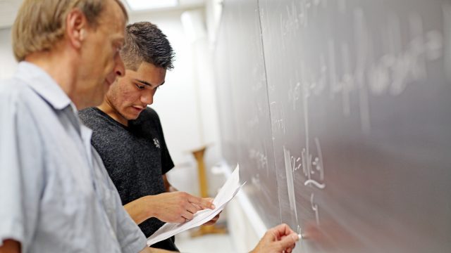 Student pointing at paper while professors writes on chalkboard