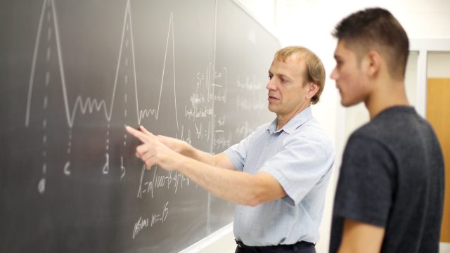 Professor pointing to chalkboard with math equations on it with student looking