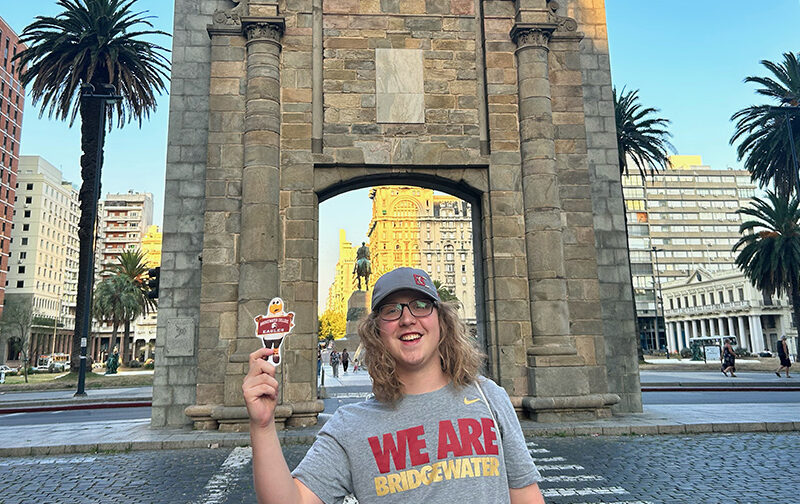 Student standing in front of the Gateway to Citadel in Uruguay in a Bridgewater College tshirt and holding flat Ernie