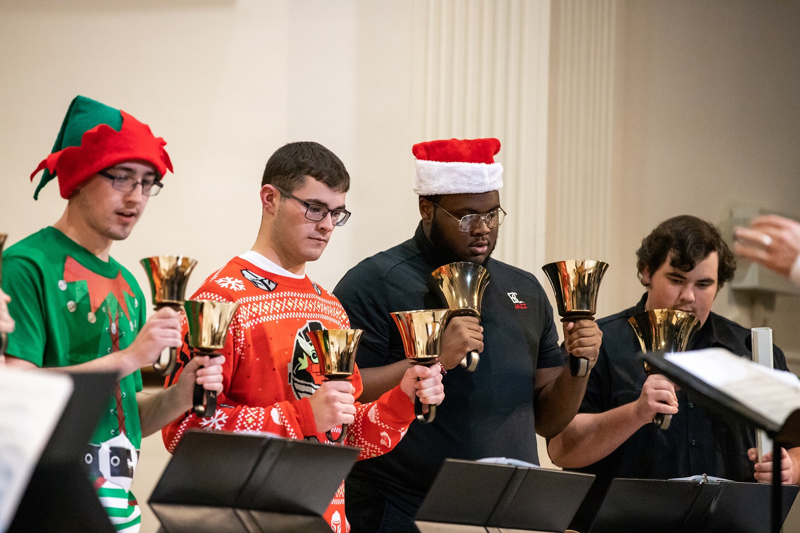 Three male students wearing holiday themed attired and holding hand bells