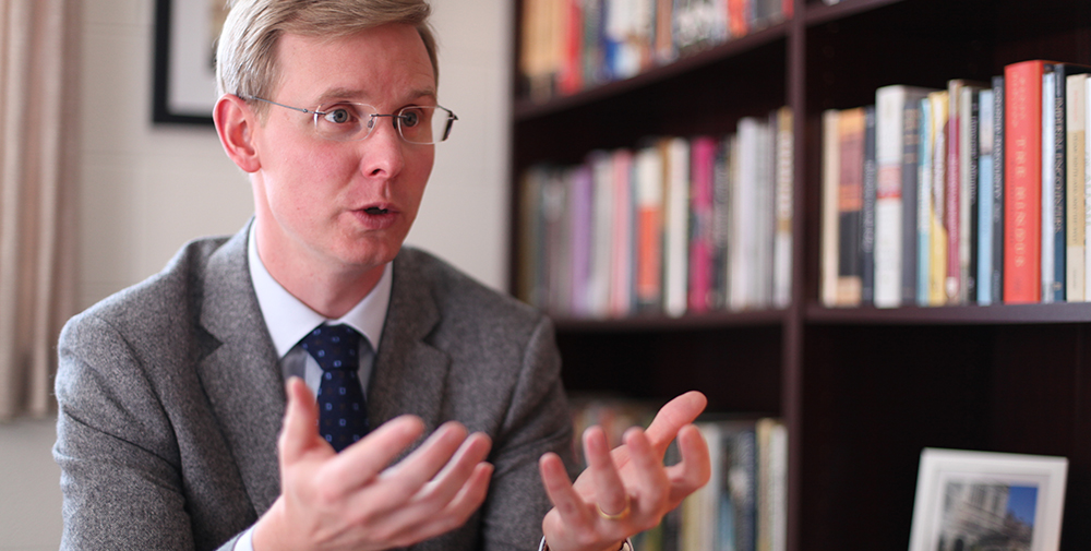 Professor speaking with hands out and a large bookshelf in the background