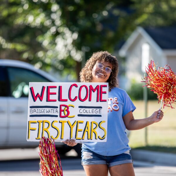 A female student holding a sign that says welcome B-C first years. In her other hand is a pompom