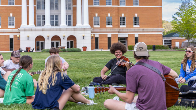 Group of students sitting a circle on the grass playing music together