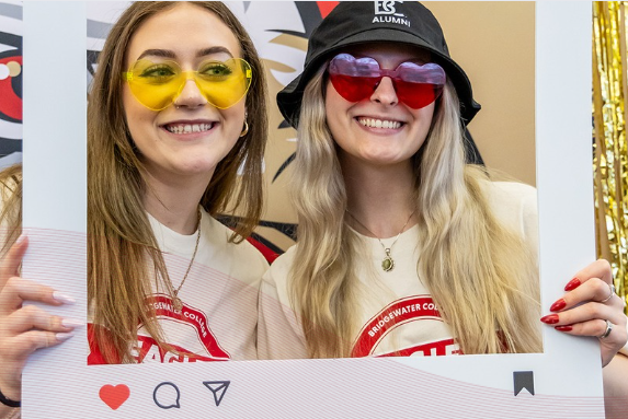 Two students wearing gold and crimson heart shaped glasses posing at the Day of Giving photobooth