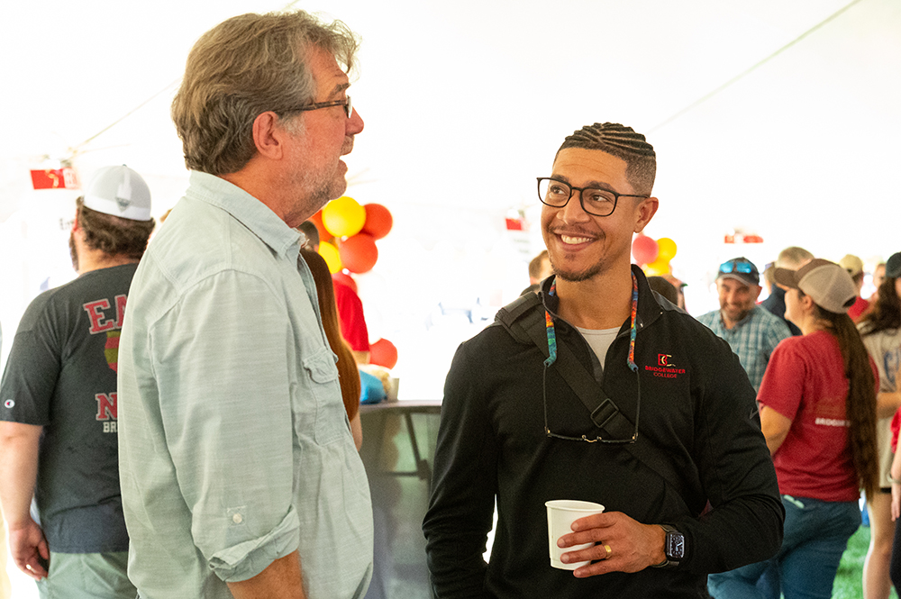 One man wearing glasses with coffee in hand talking to another man