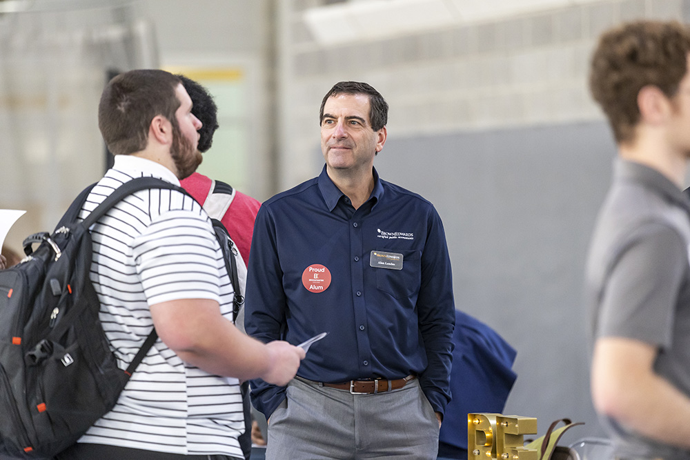 Person in blue button down shirt with proud alumni sticker talking to student at career fair