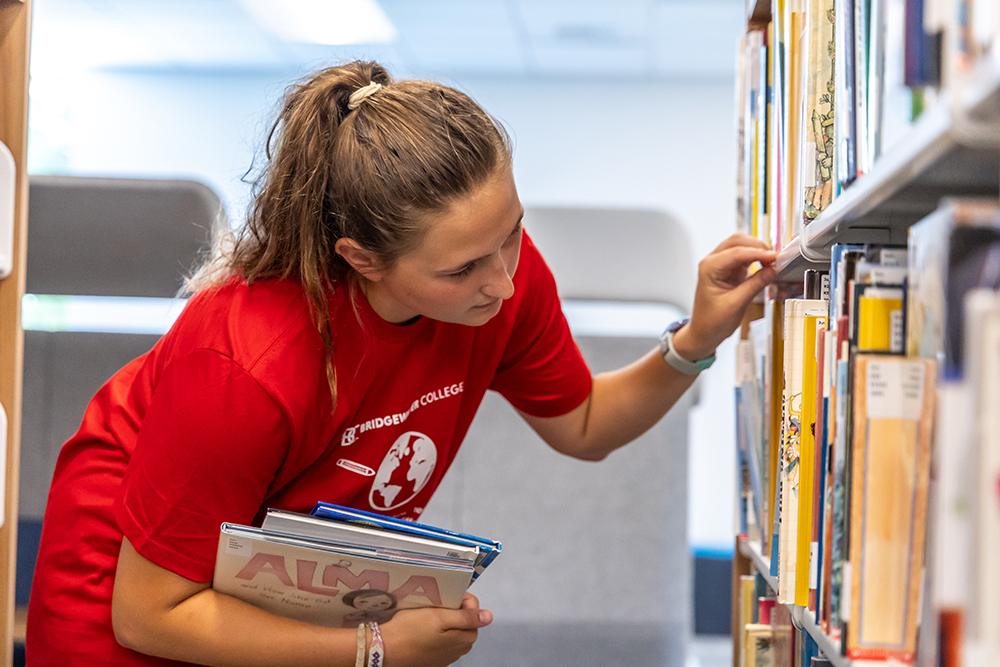 Student in red shirt holding books while looking at a shelf of books