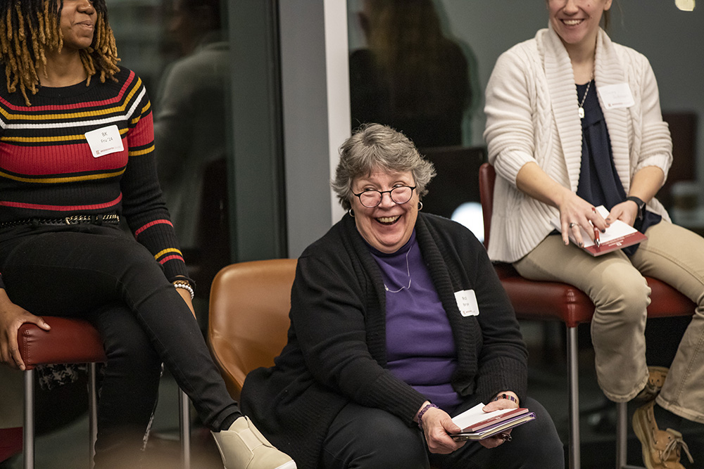 Woman wearing purple and black sitting down smiling and holding notebook