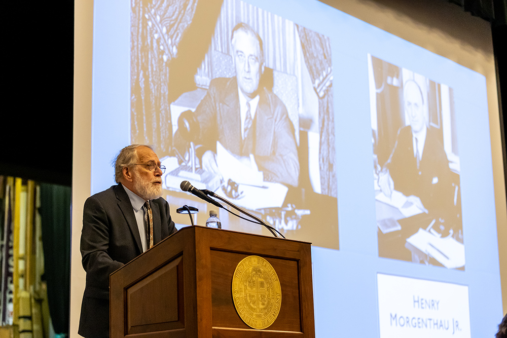 Dr. David Hollinger standing at a podium with presentation displayed on a large screen behind him