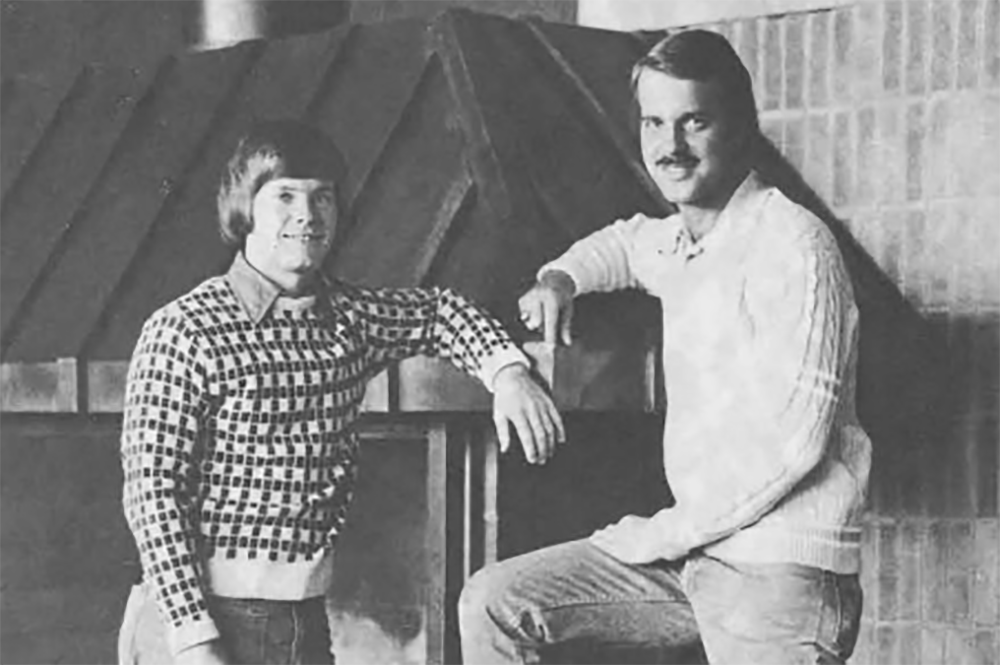 Two seniors in 1976 posing for a photo with arms rested on fireplace