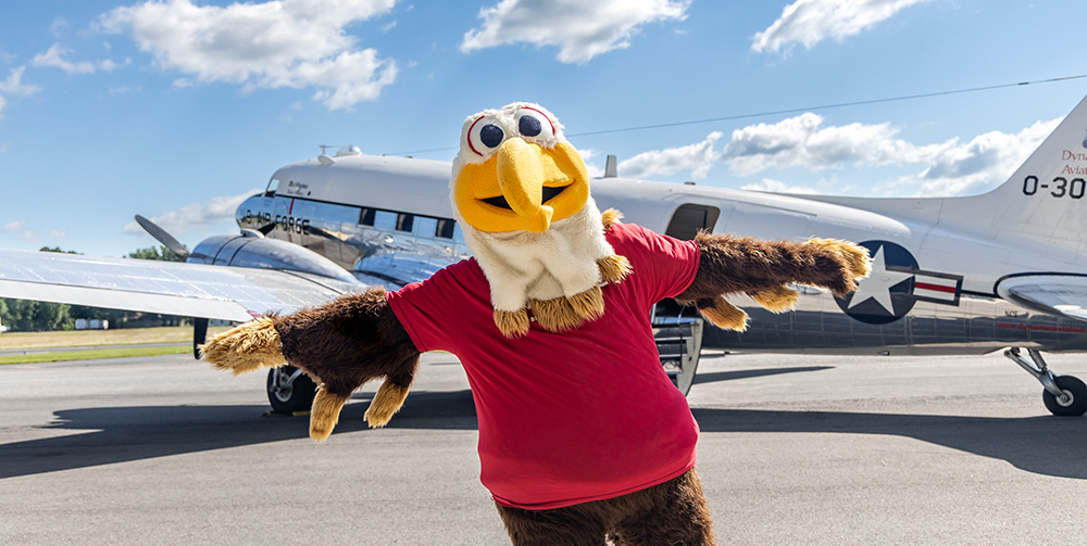 Mascot Ernie standing in front of a plane with arms spread out like a plane