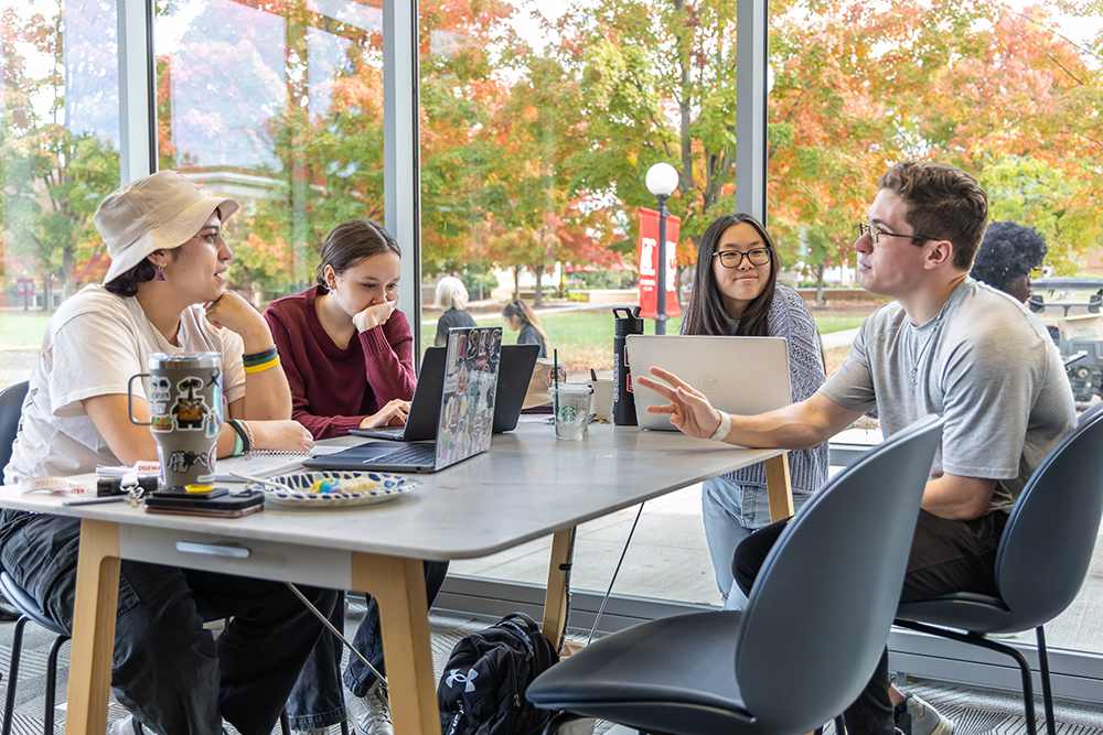 4 students sitting at a table in the library with fall leaves seen in the windows in the background