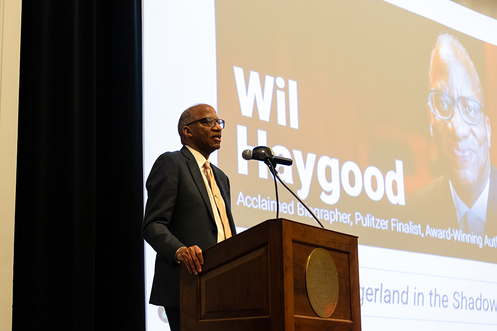 Wil Haygood speaking in front of a podium with a projector screen in the background with his name and picture