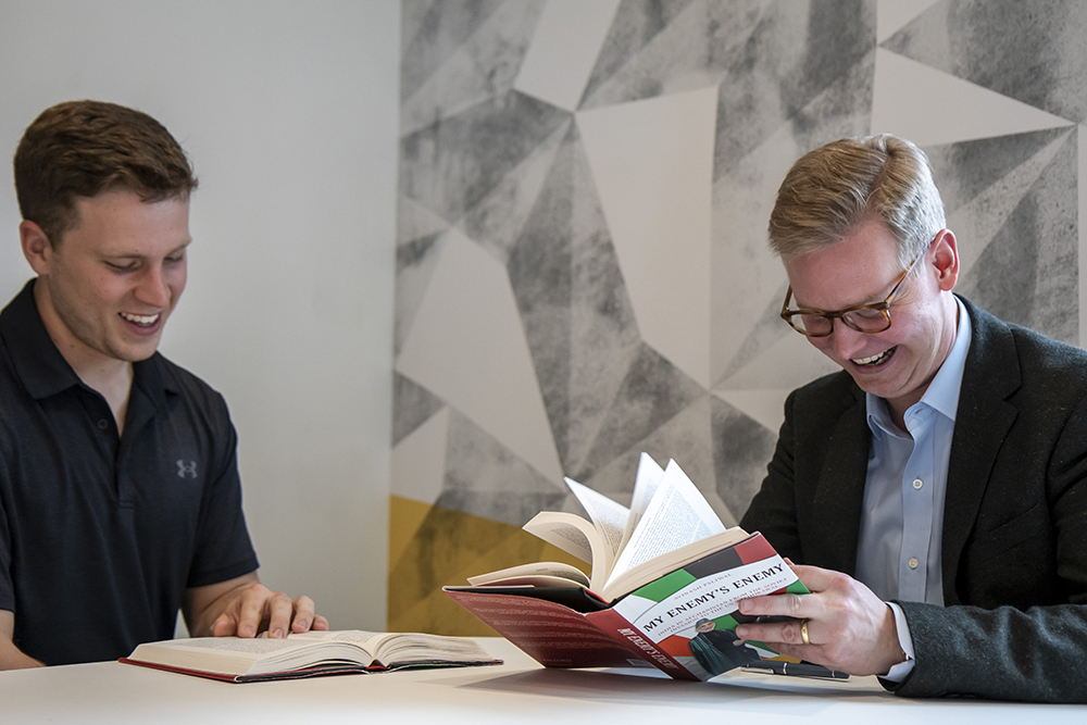 Professor Brandon Marsh meeting with a student. Both sitting at a table looking at a book and smiling. 