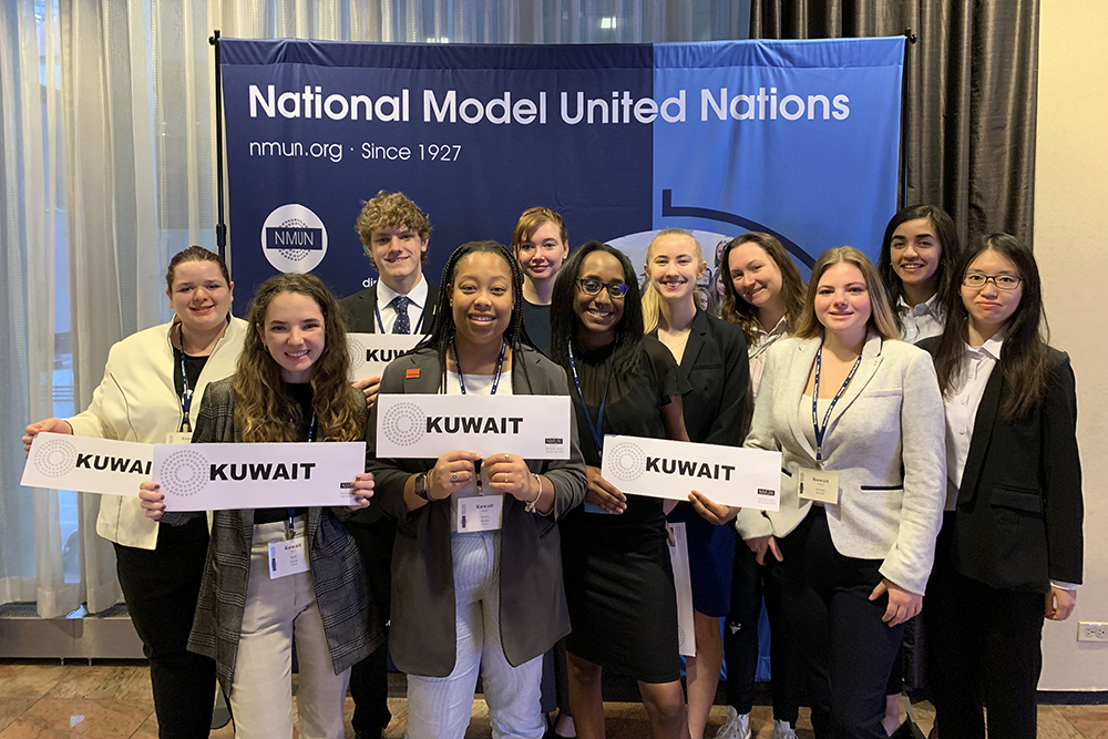 Group of students gathered in front of blue pop up banner that reads 'National Model United Nations. nmun.org since 1927" 5 students holding white sign that read 'Kuwait'