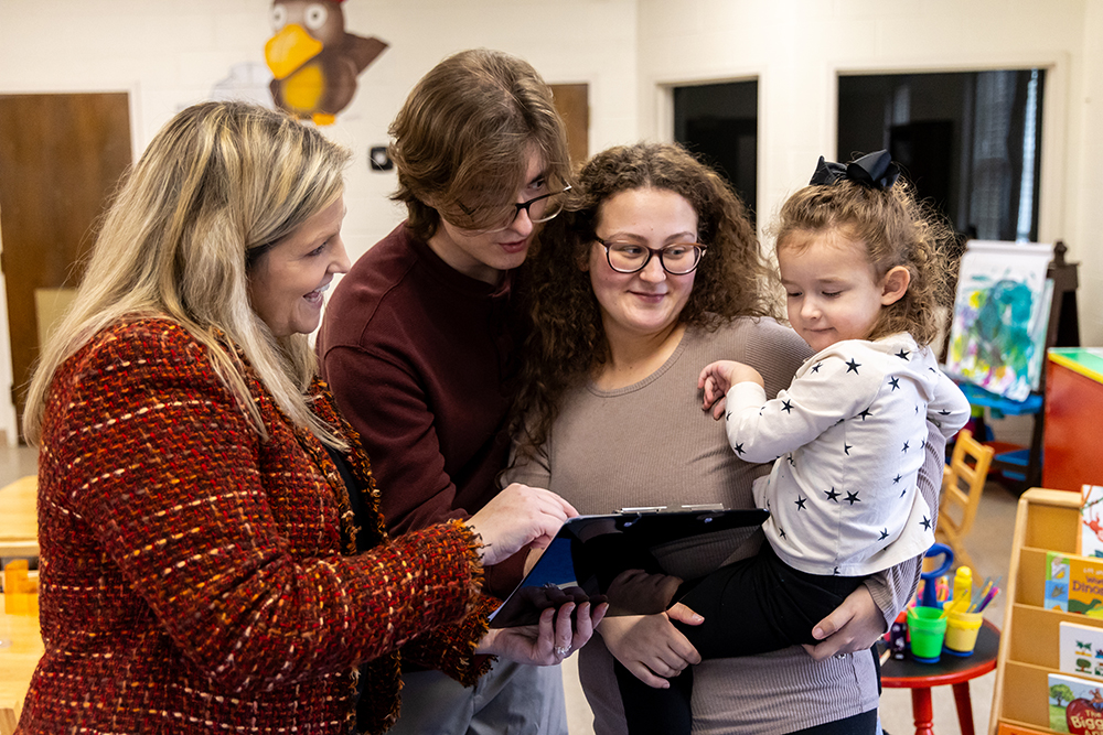 Woman pointing a black clip board to a family of a mom, dad, and daughter