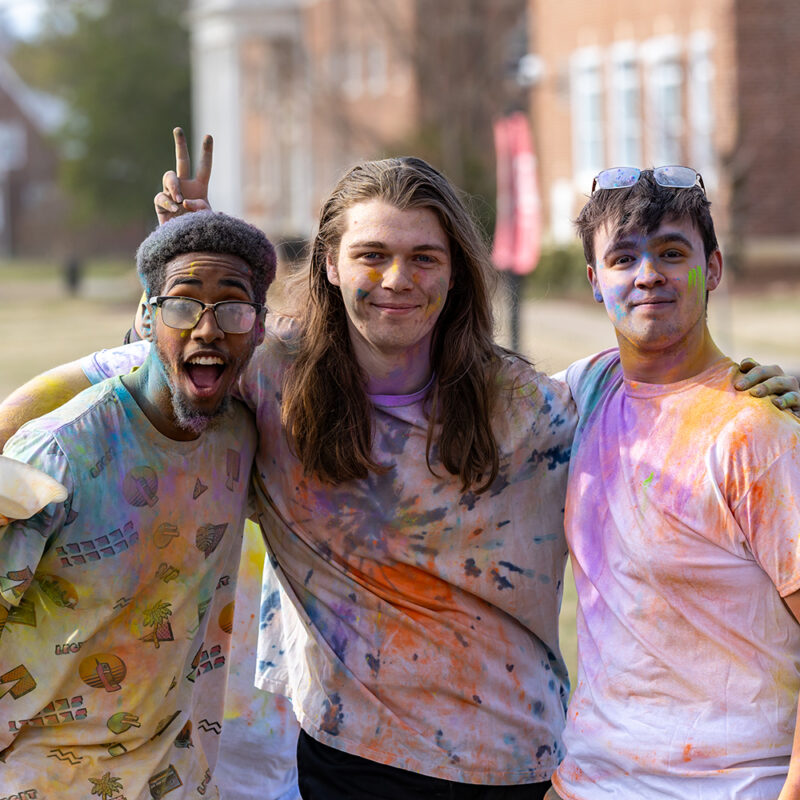 Three students covered in bright powder smiling during Holi event