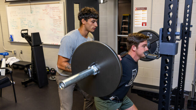 Student squating at squat rack with another student spotting